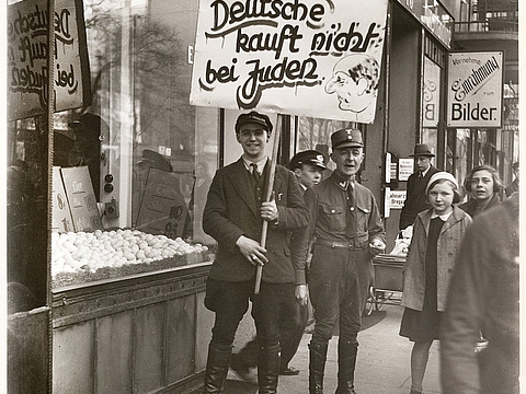 Drei Männer in Uniform stehen vor einem Geschäft. Ein Mann hält ein Schild mit der Aufschrift "Deutsche kauft nicht bei Juden" hoch. Am Rand des Fotos sind Passanten abgebildet.