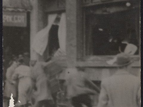 Ein Foto von dem Erdgeschoss eines Hauses mit entglaster Fensterfront. Mehrere Männer bewegen sich auf das Haus zu und werfen Gegenstände in Richtung der Fensterfront.