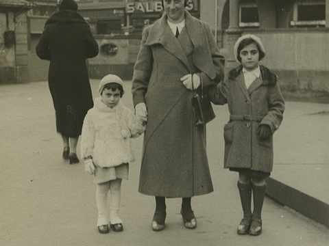Ein Foto von einer Frau, die an ihren beiden Händen junge Mädchen hält. Die Kinder stehen links und rechts von ihr. Sie alle tragen Handschuhe, Mäntel und Kopfbedeckungen und stehen auf einem Platz in einer städtischen Umgebung.