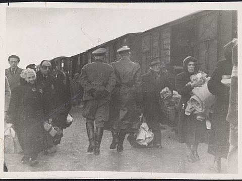 Ein Foto von zwei Männern in Mänteln, Stiefeln und Kappen die mit dem Rücken zur Kamera laufen. Links und rechts neben ihnen laufen Männer und Frauen auf die Kamera zu. Diese Menschen haben viel Gepäck auf dem Arm. Eine Frau links im Bild hat einen sechseckigen Stern auf dem Mantel aufgenäht. Im Hintergrund steht ein Güterzug.