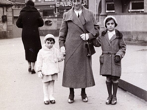 Zwei kleine Kinder stehen links und rechts von ihrer Mutter auf einem Platz vor einer Häuserfront.