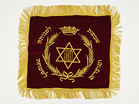 Ein Foto von einer quadratischen Decke aus dunkelrotem Samt. Auf der Decke sind ein gelber sechseckiger Stern, ein Kranz und hebräische Schriftzüge gestickt.