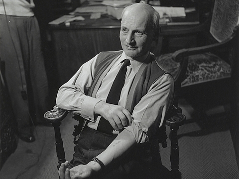 Ein Foto von einem älteren Mann, der mit Hemd und Krawatte auf einem Stuhl sitzt und seinen linken Arm leicht anhebt. Unter dem hochgekrempelten Hemd sind Schriftzeichen auf seinem Unterarm zu erkennen. Hinter dem Mann steht ein Tisch und ein weiter Stuhl und in der linken Ecke des Bildes sind die Beine eines stehenden Mannes zu erkennen.