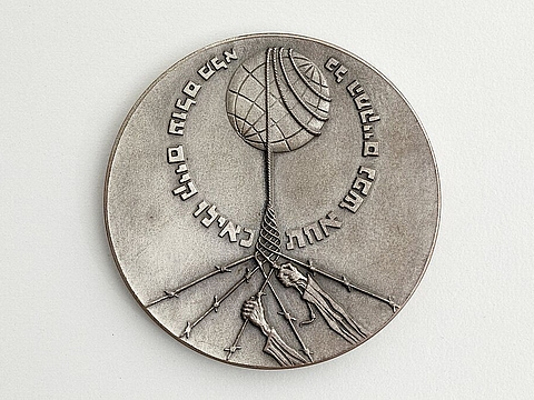 Medaille mit hebräischer Aufschrift, darauf zwei Hände an Seilen, die eine Kugel umspannen.