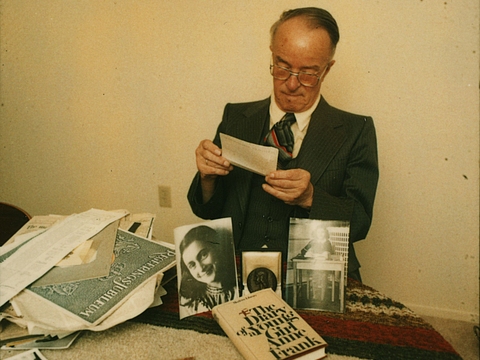Ein Foto von einem älteren Mann in Anzug und Krawatte mit Brille, der auf ein Blatt Papier blickt. Das Papier hat er in seinen Händen. Er sitzt vor einem Tisch, auf denen einige Schriftstücke und Bücher liegen. 