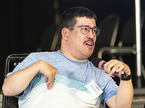 Auf dem Foto ist ein Mann zu sehen, der ein Mikrofon in der Hand hat und redet.