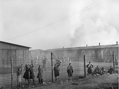 Mehrere Personen stehen an einem Zaun, hinter ihnen flache Gebäude und Rauch.