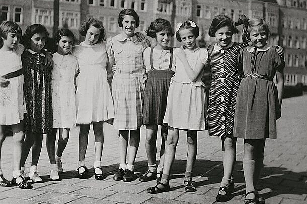 Neun Kinder Arm in Arm in Sommerkleidern auf einem Platz, dahinter hohe Wohnhäuser.