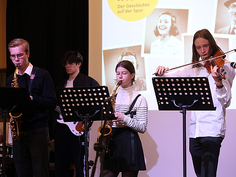 Vier Schüler*innen stehen auf einer Bühne und spielen unterschiedliche Musikinstrumente.