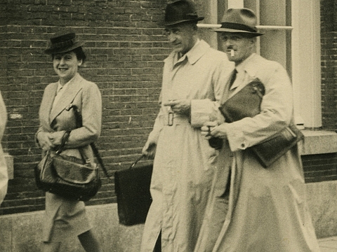 Ein Foto von einer Frau und zwei Männern. Die Drei tragen Hüte, Taschen und Mäntel und gehen vor einer Hauswand entlang.