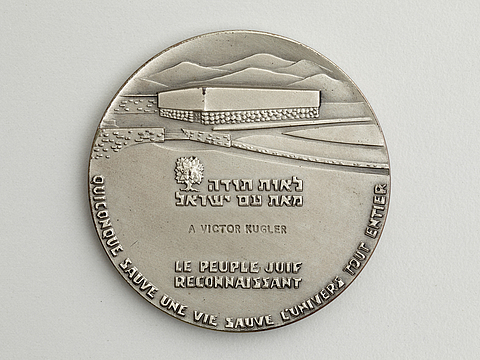 Ein Foto von einer silbernen Plakette mit Prägung. Darauf ist eine Landschaft mit Bergen und einem großen viereckigen Gebäude und Schriftzüge in Hebräisch und Französisch zu sehen. In der Mitte der Plakette steht »A Victor Kugler«.