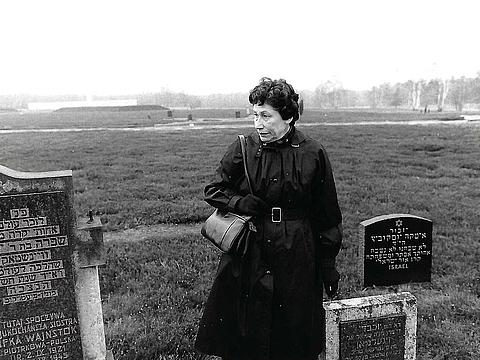 Ältere Person im Mantel auf einer Wiese neben Grabsteinen mit hebräischer Schrift.