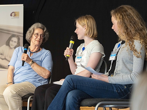 Auf dem Foto sind drei Personen zu sehen, die auf Stühlen sitzen. Links sitzt eine ältere Frau und neben ihr zwei Mädchen. 