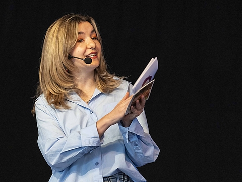 Auf dem Foto ist eine Frau zu sehen, die Karten in der Hand hat und ein Mikrofon Headseat auf dem Kopf hat. Der Hintergrund ist schwarz.
