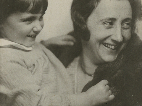 Ein Foto von einer Frau und einem jungen Mädchen. Die Frau trägt einen Mantel und hält das Mädchen auf dem Arm. Beide schauen zur Seite und lächeln.