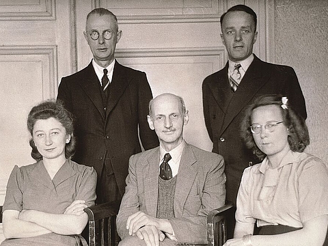 Drei Erwachsene sitzen auf Stühlen, hinter ihnen stehen zwei weitere. Alle schauen in die Kamera.