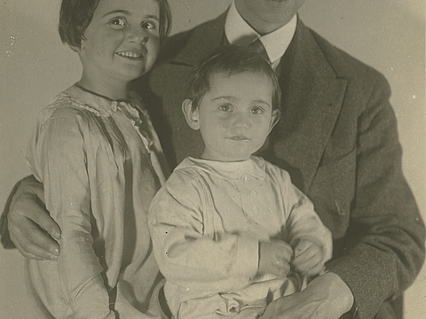Ein Foto von einem Mann im Anzug, der auf einem Stuhl sitzt und zwei junge Mädchen, die auf seinem Schoß sitzen. Die beiden Mädchen schauen in die Kamera und der Mann schaut auf das kleinere Mädchen herab.