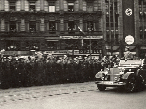 Ein Foto von einem fahrenden Auto mit geöffnetem Verdeck, in dem ein Mann steht und auf eine jubelnde Menschenmenge blickt. Die Häuserfront im Hintergrund trägt ein langes Banner mit einem Hakenkreuz drauf.