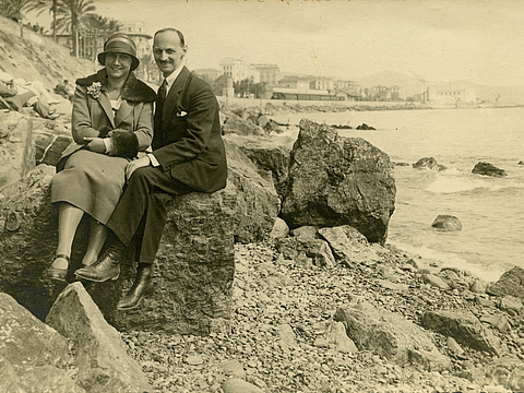 Ein Foto von einer Mann und einer Frau, die nebeneinander auf einem großen Stein sitzen und lächeln. Im Hintergrund ist Wasser, Häuser und Palmen zu sehen. Neben ihnen sind noch mehr Steine zu sehen. Die Frau trägt einen Hut und der Mann einen Anzug.