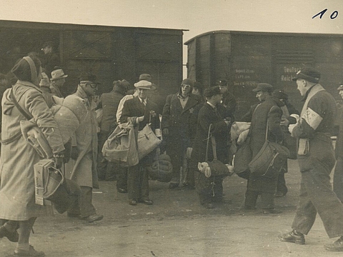 Menschengruppe in Winterkleindung und mit Taschen, dahinter zwei Viehwaggons.