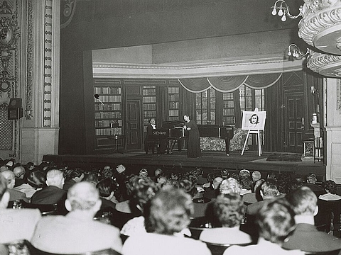 Viele Menschen sitzen vor einer Bühne, auf der jemand am Klavier spielt und eine Frau singt.