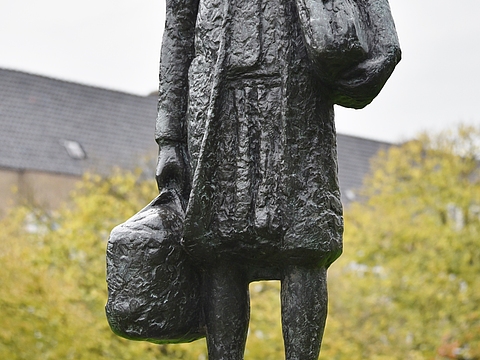 Ein Foto von einer dunklen Statue eines jungen Mädchens mit zwei Taschen in den Händen. Die Statue steht auf braunem Stein. Auf dem Stein steht in weißer Schrift »Anne Frank« geschrieben, im Hintergrund ist ein Haus zu sehen.