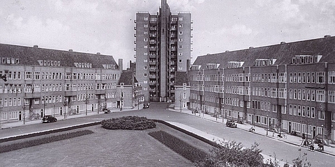 Platz mit Grünfläche, dahinter vierstöckige Wohnhäuser. Hochhaus im Hintergrund.