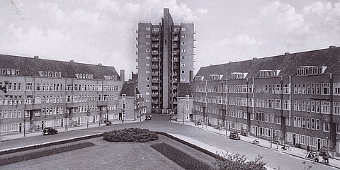 Platz mit Grünfläche, dahinter vierstöckige Wohnhäuser. Hochhaus im Hintergrund.