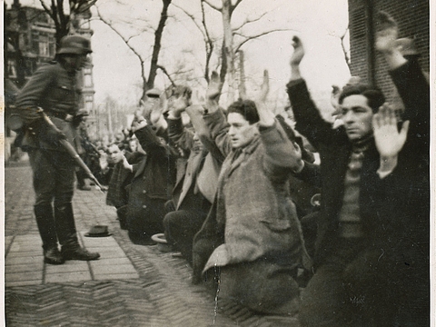 Mehrere Reihen Männer knien auf dem Boden und strecken ihre Arme in die Luft. Ein bewaffneter Polizist steht vor ihnen.