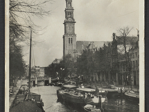 Ein Foto von einem Kanal zwischen Häusern. Im Wasser liegen Schiffe vor Anker und ein großes Schiff fährt auf dem Kanal. Im Hintergrund ist ein hoher Kirchturm zu sehen und auf den Straßen neben dem Kanal sind Bäume ohne Blätter.
