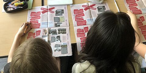 Auf dem Foto sind zwei Mädchen zu sehen, die auf eine Zeitung schauen. Die Zeitung liegt auf einem Tisch. Der Hinterkopf der beiden Mädchen ist zu sehen.