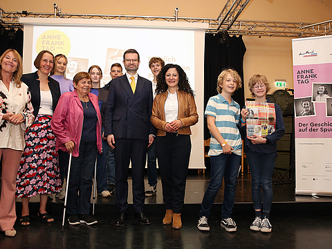Das Foto zeigt einige Menschen verschiedenen Alters auf einer Bühne. Sie lächeln in die Kamera. Rechts von Ihnen steht ein Bildaufsteller mit einem Ausstellungsmotiv.