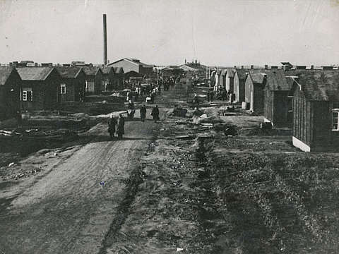 Ein Foto zeigt links und rechts Häuserbaracken. Dazwischen ein Weg, auf dem Menschen laufen. Im Hintergrund ein großer, schmaler Schornstein.