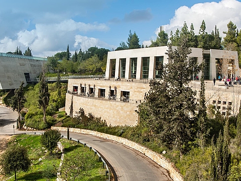 Ein großes längliches Gebäude mit vielen Fenstern steht an einer Straße zwischen einigen Bäumen.