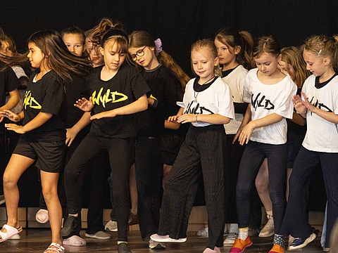 Auf dem Foto sind viele Kinder zu sehen, die in Bewegung sind. Alle tragen gleiche T-Shirts, die auf der rechten Seite haben weiße Oberteile an und die Personen auf der linken Seite schwarze.