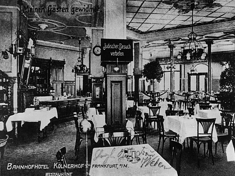 Ein Foto von einem Saal, in dem viele Tische stehen, die mit Geschirr gedeckt sind und Tischdecken haben. An den Tischen stehen Stühle. In der Mitte des Raums steht eine Säule mit einem Schild, auf dem »Jüdischer Besuch verbeten« steht.