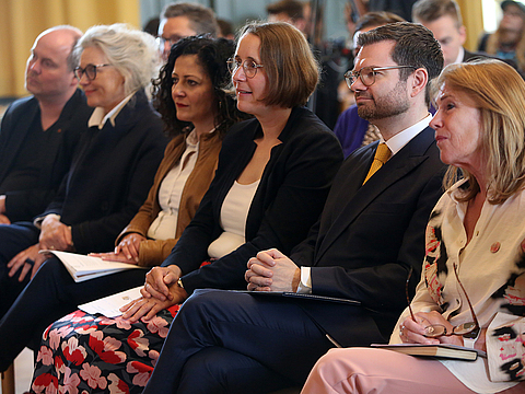 Sechs Menschen sitzen nebeneinander in der ersten Reihe eines Publikums.