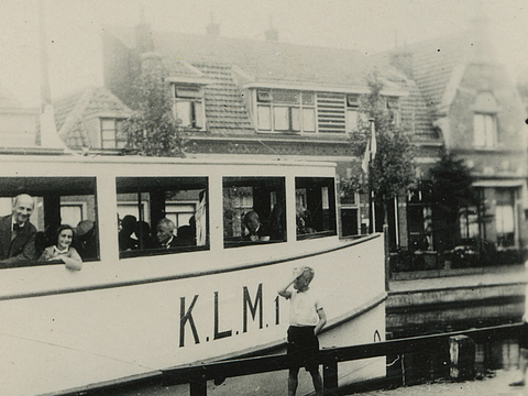 Ein Foto von einem Teil eines Boots auf einem Kanal, aus ein Mädchen und ein Mann gucken. Im Hintergrund ist eine Häuserfront zu sehen.