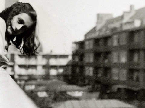 Ein Mädchen lehnt an einem Balkongeländer und blickt in die Kamera. Im Hintergrund stehen Häuser.