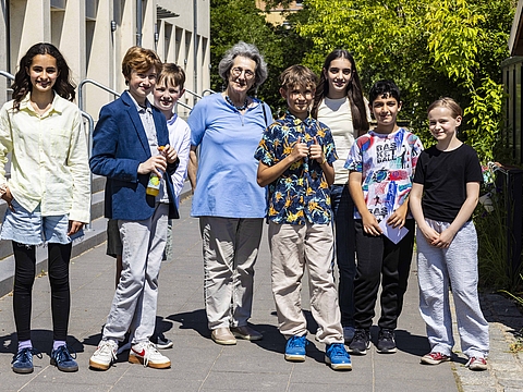 Draußen vor einem Gebäide stehen sieben junge Menschen und in ihrer Mitte steht eine ältere Frau mit grauen Haaren. Alle Menschen lächeln in die Kamera und sind auch zu ihr positioniert.