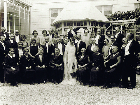 Viele Menschen in feierlicher Kleidung stehen links und rechts von einem Brautpaar. Im Hintergrund steht ein Haus mit Wintergarten.