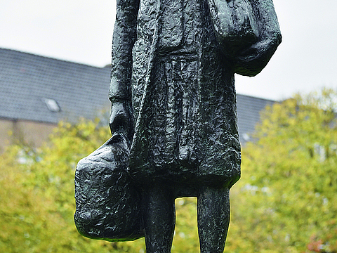 Eine dunkle Statue von einem jungen Mädchen mit zwei Taschen in den Händen steht auf einem braunen Stein. Auf dem Stein steht in weißer Schrift »Anne Frank« geschrieben, im Hintergrund ist ein Haus zu sehen.