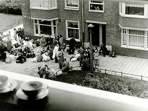 Blick aus einem Fenster auf eine Straße, darauf eine Gruppe von Menschen mit Gepäck.