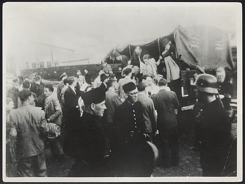 Ein Foto von einer Gruppe uniformierter Männer im Vordergrund, die einander zugewandt stehen. Im Hintergrund steht ein Kraftwagen, aus dem zivil gekleidete Menschen von der Ladefläche heruntersteigen. Um das Fahrzeug und neben den uniformierten Männern herum stehen weitere zivil gekleidete Menschen. 