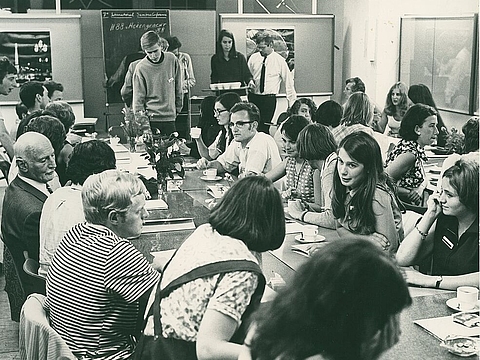 Eine ältere Person in Anzug mit jungen Menschen im Gespräch an einem langen Tisch.