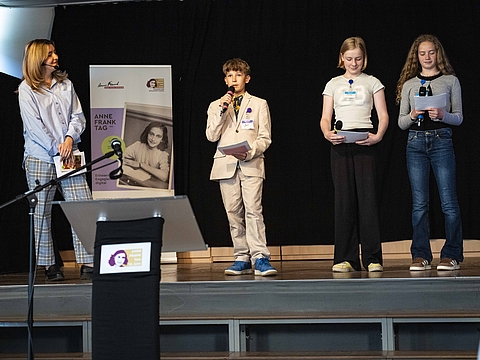 Auf dem Foto sieht man eine Bühne, auf der auf der rechten Seite drei junge Menschen stehen, die alle ein Mikrofon in der Hand haben. Links neben den Kindern ist ein Banner und links neben dem Banner steht eine Frau, die den Kindern zugewandt ist. 