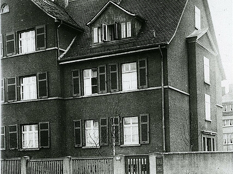 Ein zweistöckiges Mehrfamilienhaus mit Giebeldach, Fensterläden und Vorgarten.