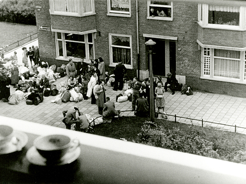 Ein Foto von einem Haus, vor dem viele Menschen, die Gepäck bei sich haben, sitzen und stehen. Im Vordergrund sind zwei Tassen mit Untertassen auf einer Fensterbank zu sehen. Das Foto ist aus einem höheren Stockwerk aufgenommen worden.
