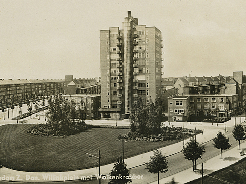 Ein Foto von einem Häuserblock mit einem großen, mehrgeschossigen Haus an der Front. Vor den Häusern ist ein Platz mit Wiesen und Bäumen umringt von einer Straße.
