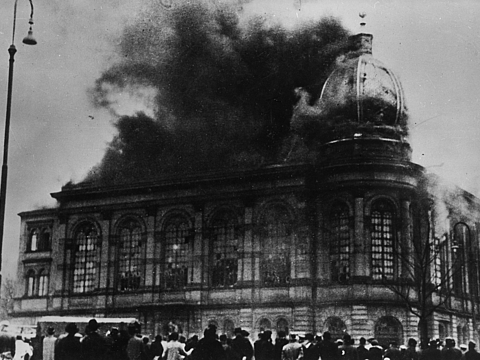 Ein Foto von einem großen Gebäude mit vielen Fenstern und mit einer Kuppel aus dem Rauch in den Himmel aufsteigt. Vor dem Gebäude stehen viele Menschen und schauen auf das Gebäude.